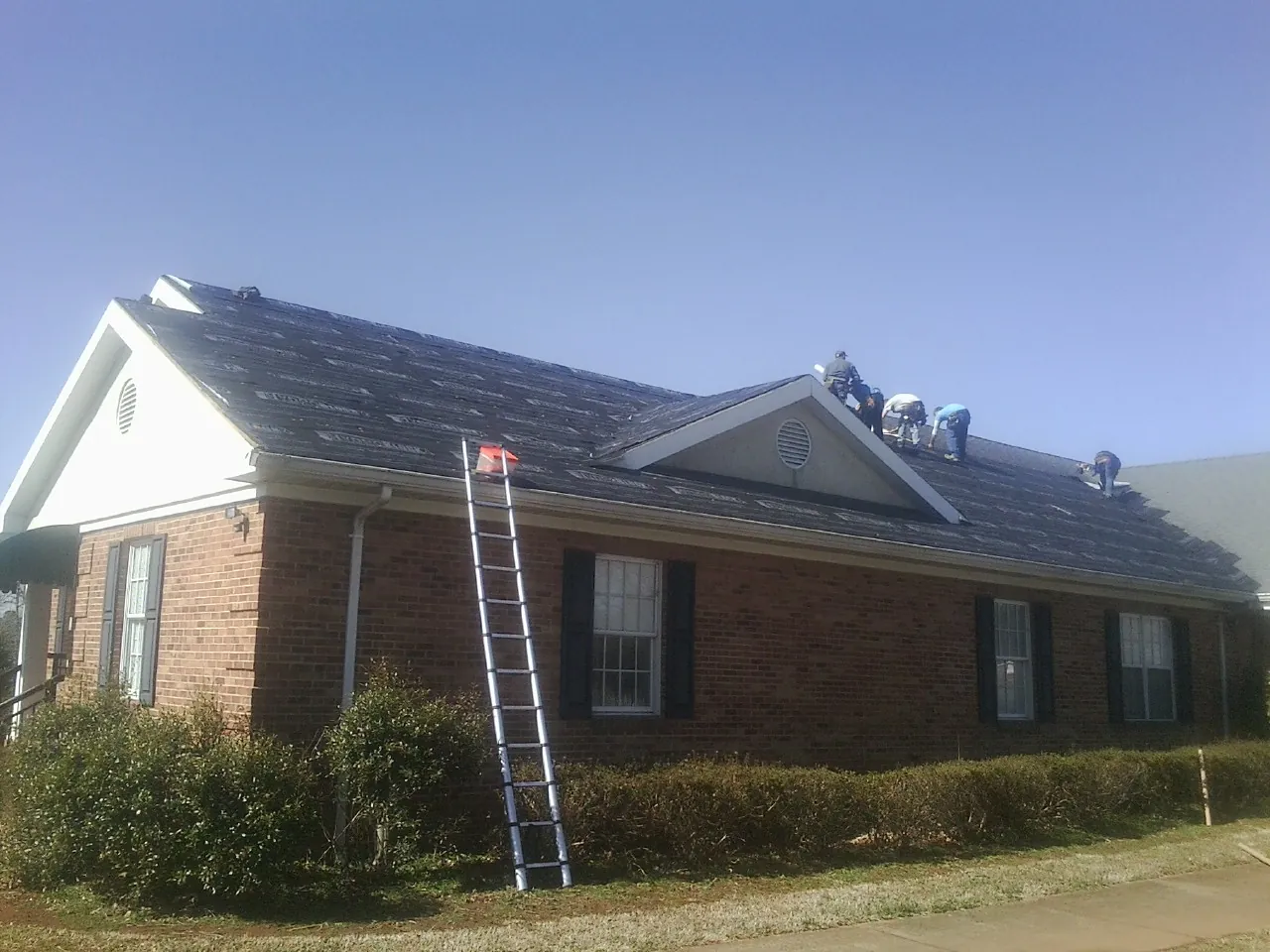 Crew tearing shingles off a brick ranch roof with a ladder set — Metal Roof Repair work by our crew in Cannonville