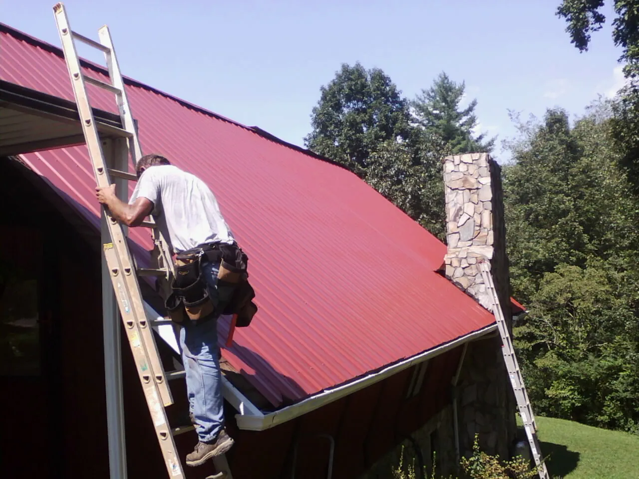 Roofer on a ladder beside a red metal roof and stone chimney — Soffit & Fascia Repair by Roofing Brothers in Cannonville